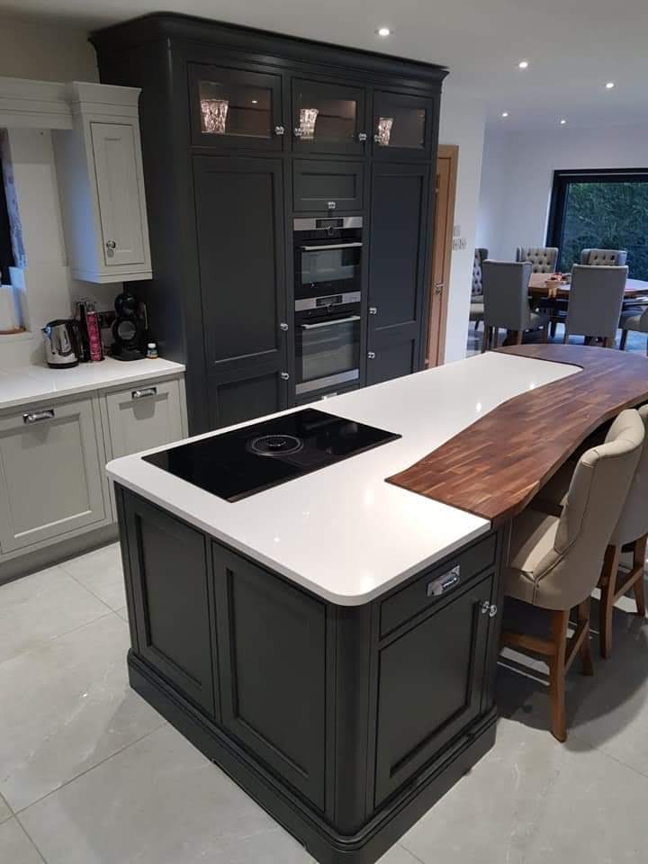 Nowoczesna kuchnia z wyspą, ciemnoszare szafki z przeszklonymi górnymi frontami, biały blat wyspy z wbudowaną płytą indukcyjną i drewnianym blatem barowym, widoczne krzesła barowe i stół jadalny...