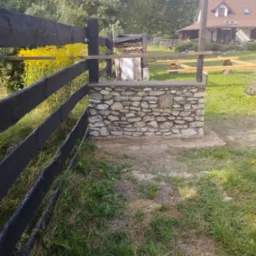 Kamienny murek, prawdopodobnie fragment ogrodzenia, w trakcie budowy; widoczny czarny drewniany płot i dom w tle.