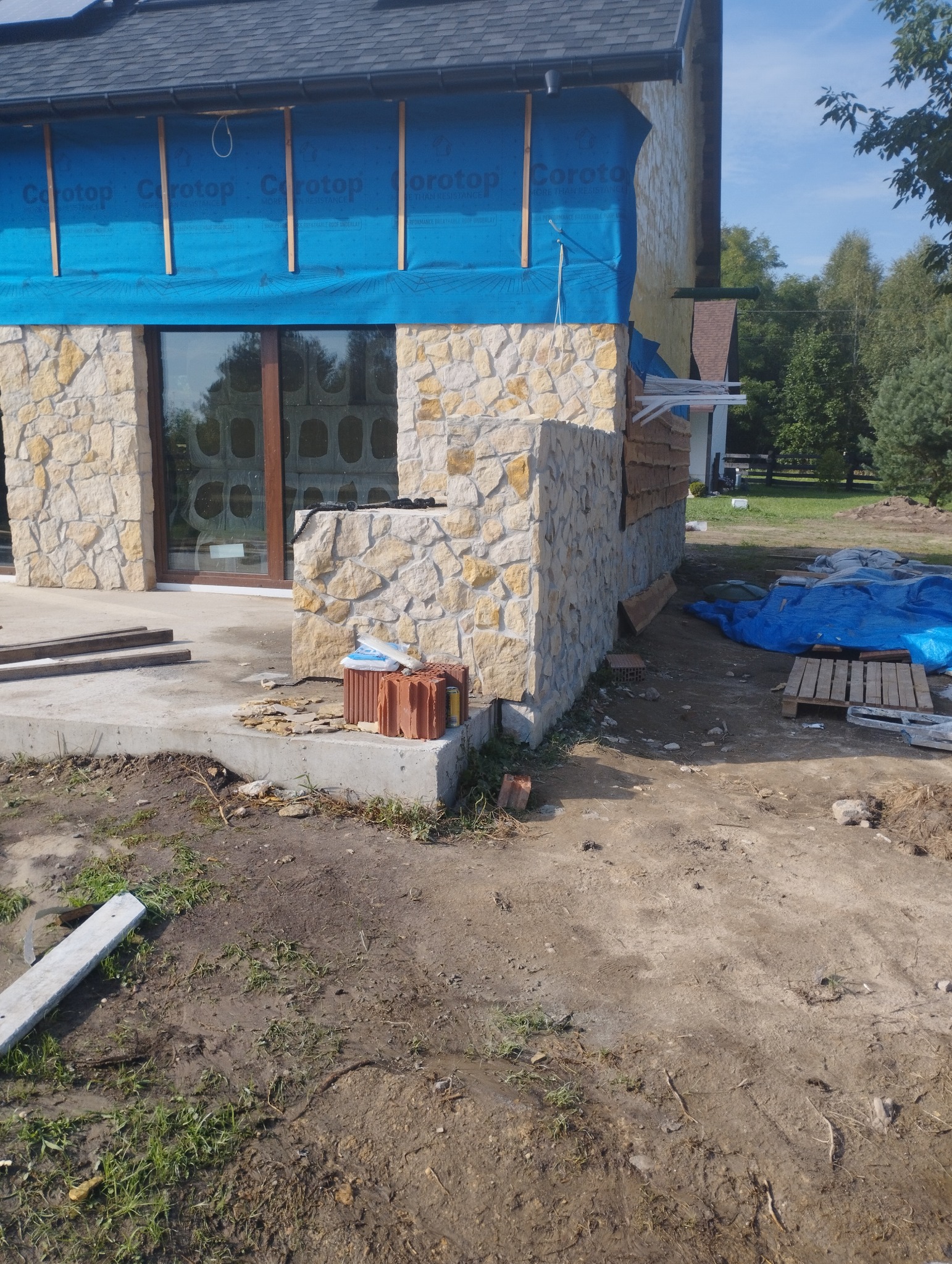 Wznoszenie ściany z kamienia naturalnego na budynku w trakcie budowy, widoczne materiały izolacyjne i cegły na fundamencie.