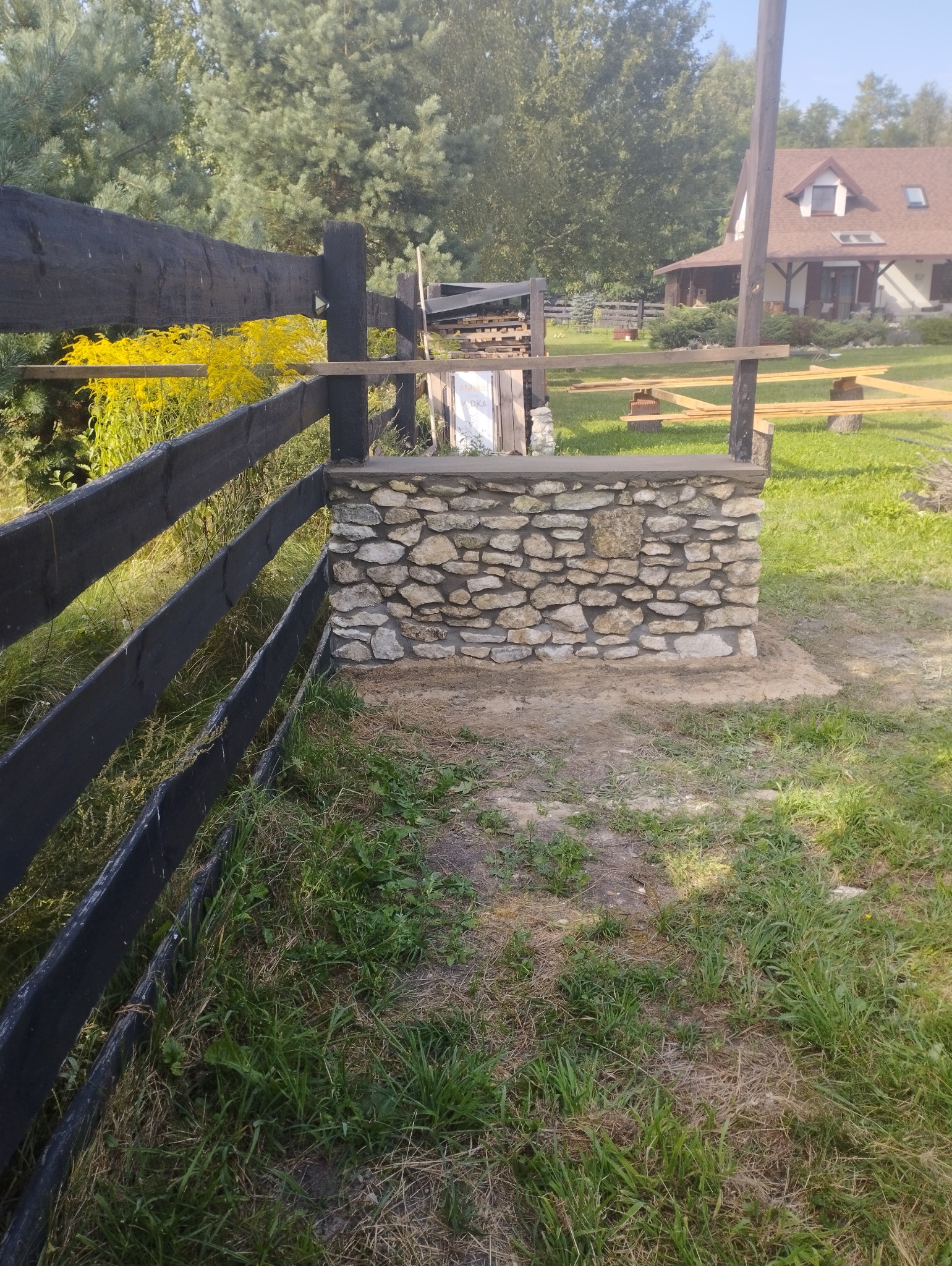 Kamienny murek, prawdopodobnie fragment ogrodzenia, w trakcie budowy; widoczny czarny drewniany płot i dom w tle.