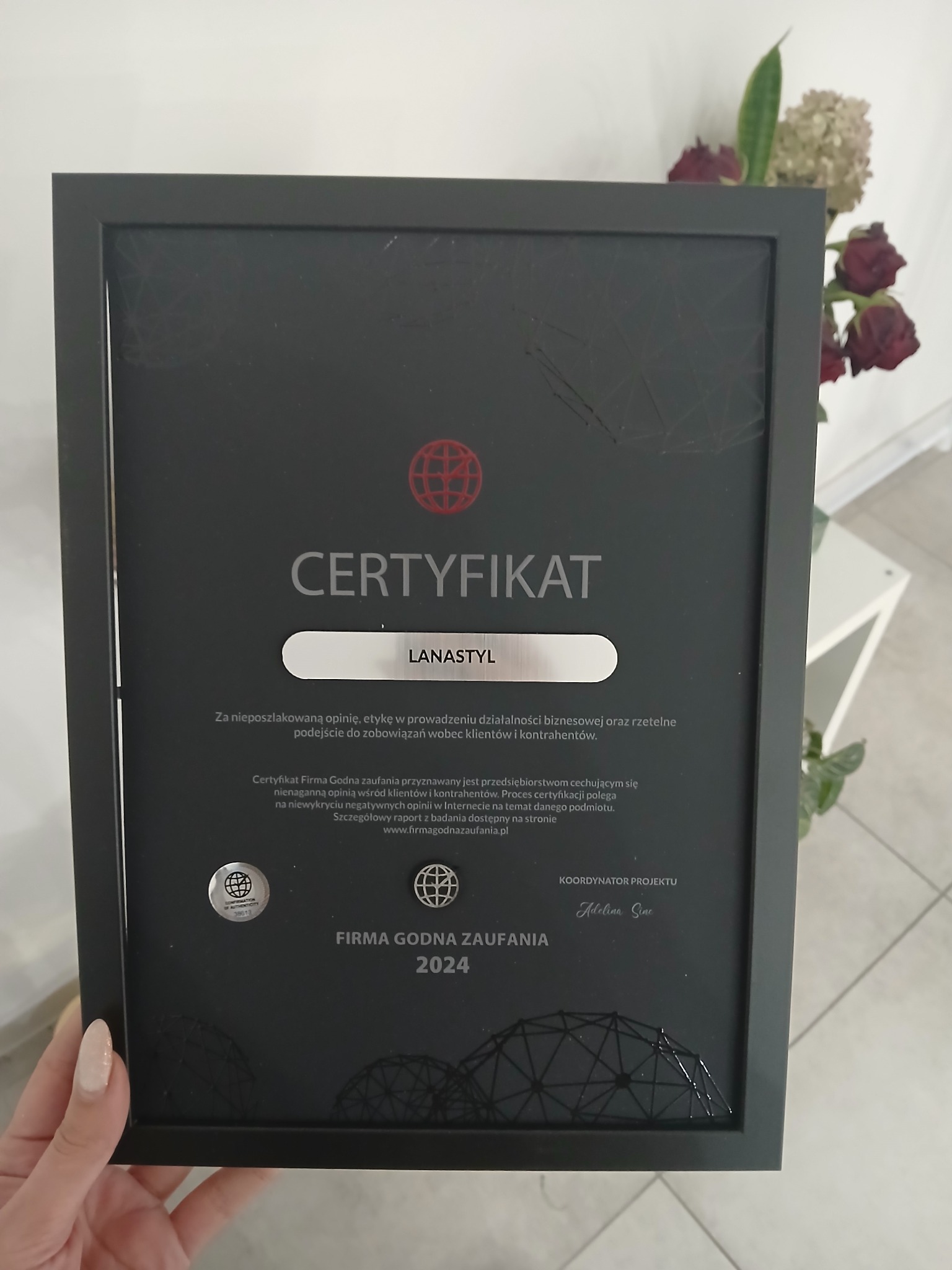 Oprawiony certyfikat 'Firma Godna Zaufania 2024' dla firmy Lanastyl, z widocznym logo i podpisem koordynatora projektu, umieszczony obok kwiatów.