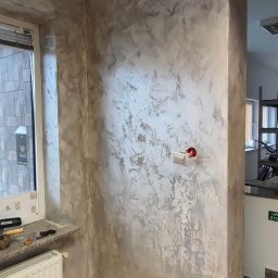 PAW-BUD Paweł Stopa - Ściana pokryta dekoracyjną, srebrzysto-szarą fakturą, widoczne gniazdko elektryczne i fragment okna z roletą, podłoga częściowo zabrudzona po remoncie.