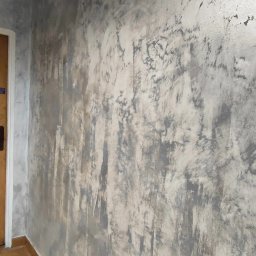 PAW-BUD Paweł Stopa - Ściana w odcieniach szarości i bieli z efektem dekoracyjnym, widoczny narożnik pomieszczenia i wiadro z narzędziami malarskimi w dolnym rogu.