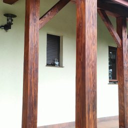 PAW-BUD Paweł Stopa - Drewniana konstrukcja z belek i słupów, zabezpieczona ciemnobrązowym lakierem, tworząca zadaszenie nad tarasem z terakotową podłogą i fragmentami roślinności.