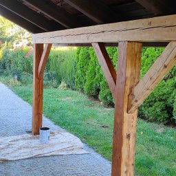 PAW-BUD Paweł Stopa - Drewniana konstrukcja zadaszenia, częściowo pomalowana bejcą, z widocznym wiadrem farby i rozłożoną folią ochronną na kostce brukowej, w tle zielony trawnik i krzewy.