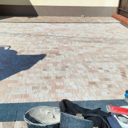 PAW-BUD Paweł Stopa - Układanie kostki brukowej na posesji: pracownik zagęszcza podłoże ubijarką mechaniczną, widoczne ułożone fragmenty w kolorze beżowym i czarnym, narzędzia pracy, poziomica, wiaderka,...