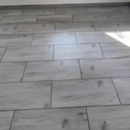 Ułożone jasnoszare płytki podłogowe imitujące drewno, z ciemnoszarymi fugami, widoczne przy ścianie listwy przypodłogowe, jasny punkt od światła słonecznego.