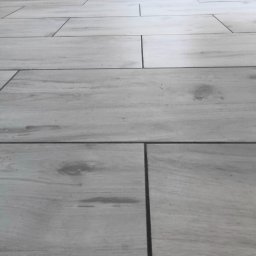 PAW-BUD Paweł Stopa - Ułożone jasnoszare panele podłogowe z widocznym wzorem drewna i ciemnymi fugami, przy białej ścianie z listwą przypodłogową z płytek.