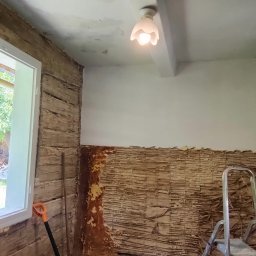 PAW-BUD Paweł Stopa - Pokój w Przeworsku w trakcie prac remontowych: odsłonięta ściana z drewnianą konstrukcją, fragment uszkodzonego sufitu, gruz na podłodze, drabina i narzędzia.