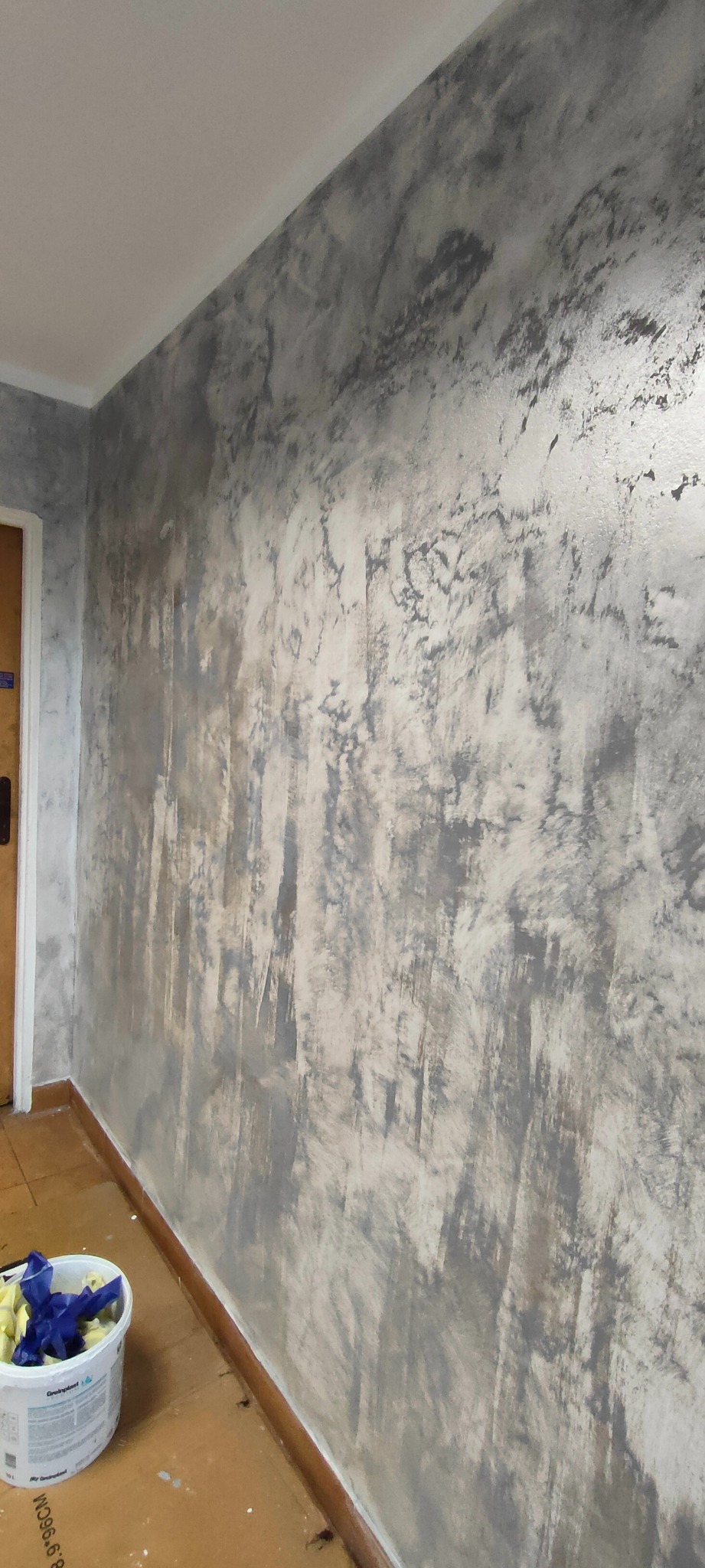 Ściana w odcieniach szarości i bieli z efektem dekoracyjnym, widoczny narożnik pomieszczenia i wiadro z narzędziami malarskimi w dolnym rogu.