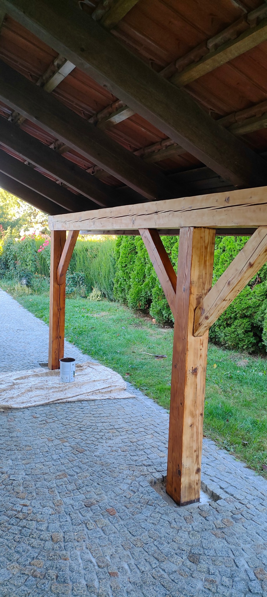 Drewniana konstrukcja zadaszenia, częściowo pomalowana bejcą, z widocznym wiadrem farby i rozłożoną folią ochronną na kostce brukowej, w tle zielony trawnik i krzewy.