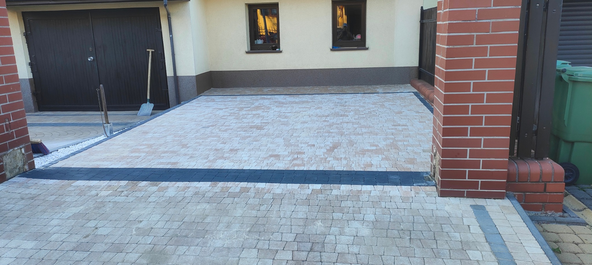 Ułożona kostka brukowa na podjeździe przed garażem, z widocznymi obrzeżami, kolumną z czerwonej cegły i narzędziami w tle.