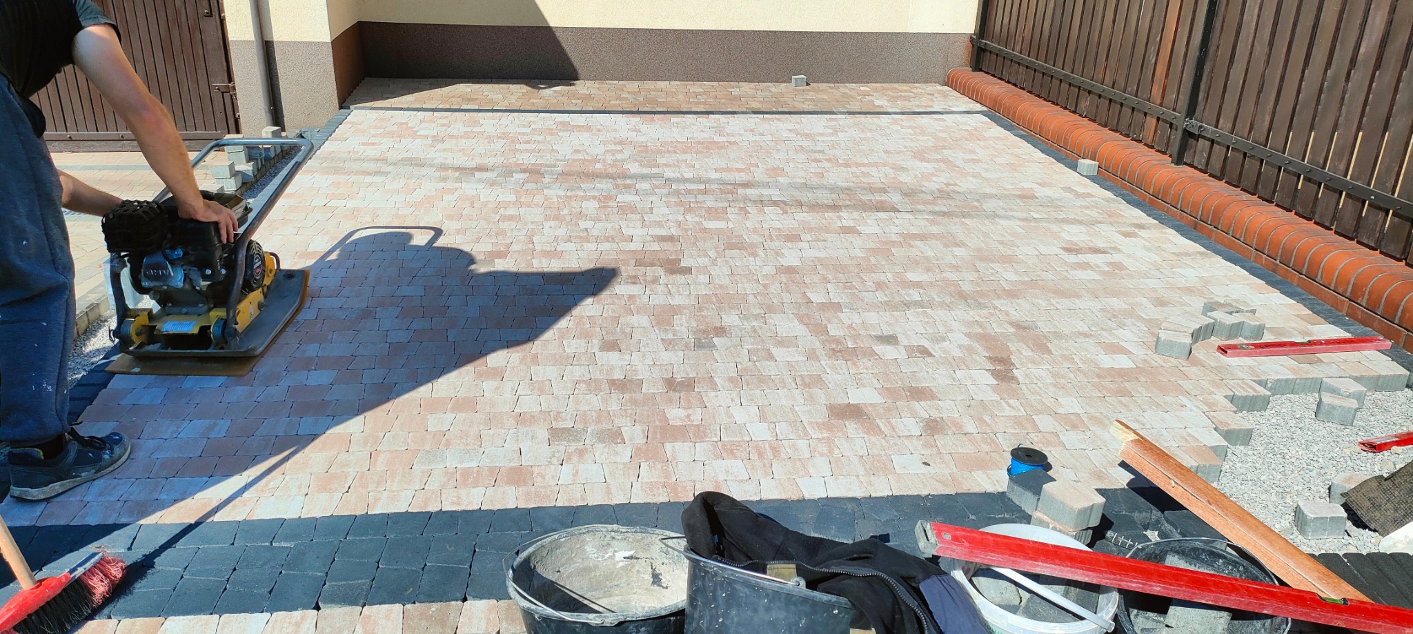 Układanie kostki brukowej na posesji: pracownik zagęszcza podłoże ubijarką mechaniczną, widoczne ułożone fragmenty w kolorze beżowym i czarnym, narzędzia pracy, poziomica, wiaderka,...