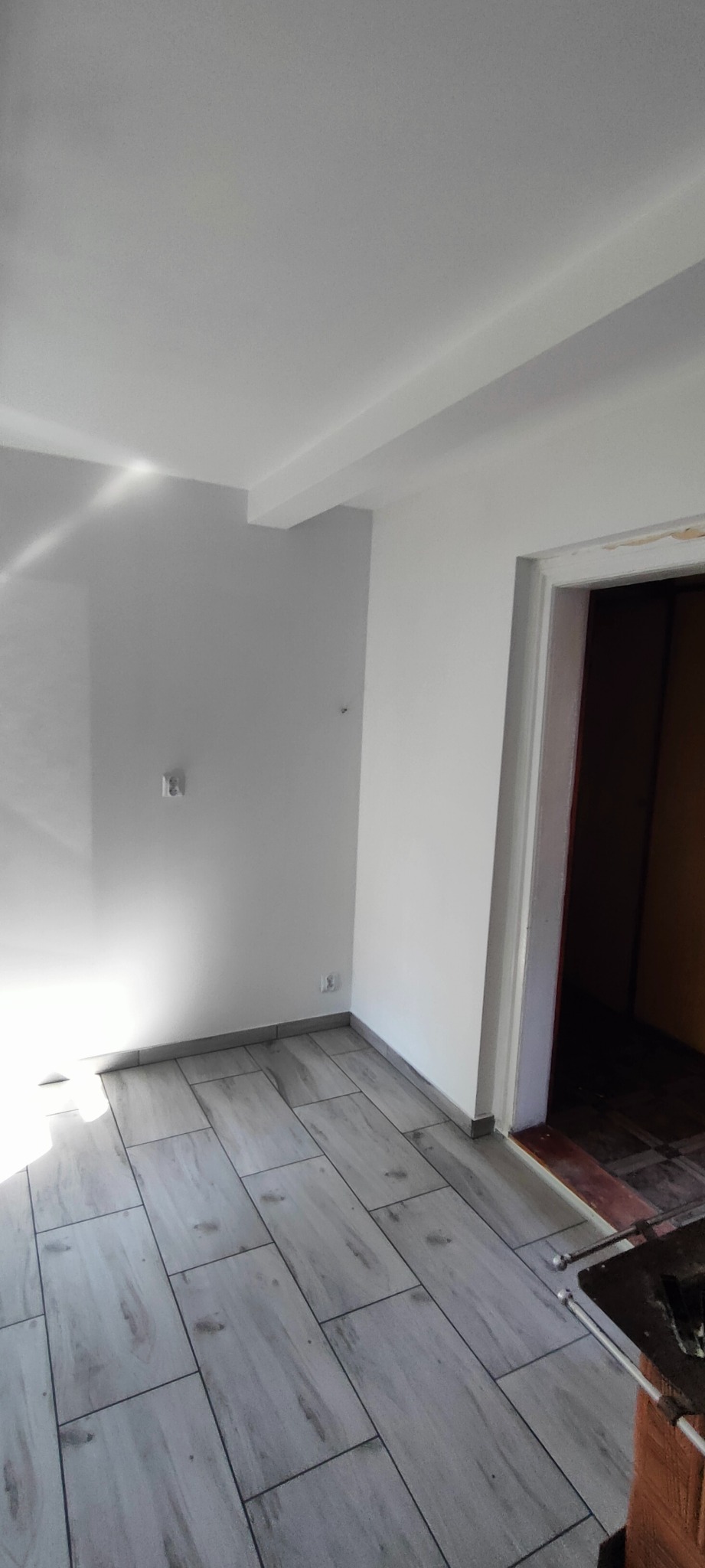 Pomalowane na biało wnętrze pokoju z szarymi panelami podłogowymi imitującymi drewno, widoczne gniazdka elektryczne na ścianie i fragment futryny drzwiowej.
