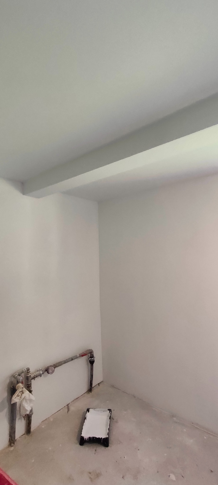 Świeżo pomalowane na biało pomieszczenie z rurami przy podłodze i kuwetą malarską z resztkami farby, widoczne niedoskonałości na podłodze.