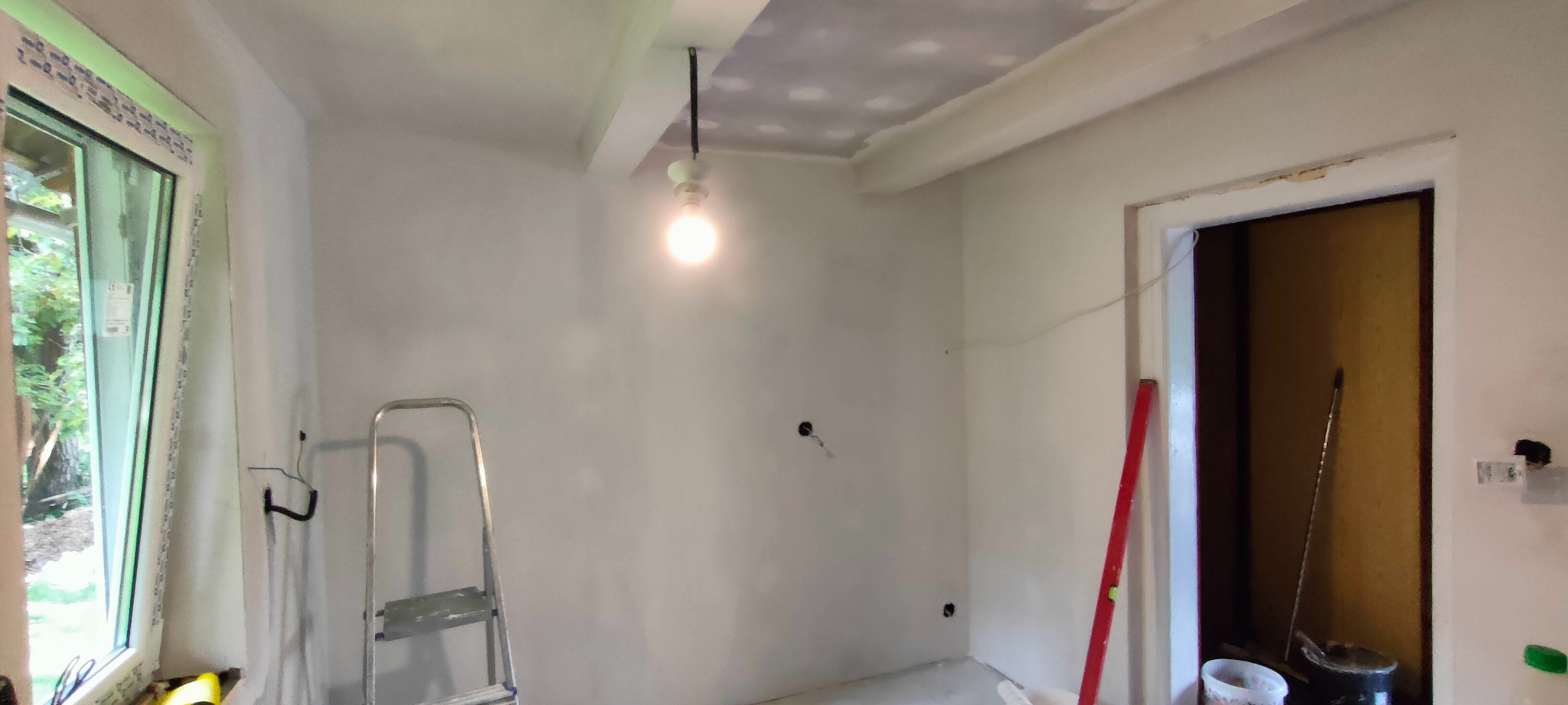 Pokój w trakcie prac przygotowawczych: ściany i sufit po nałożeniu gładzi, widoczna drabina, poziomica i prowizoryczne oświetlenie żarówką.