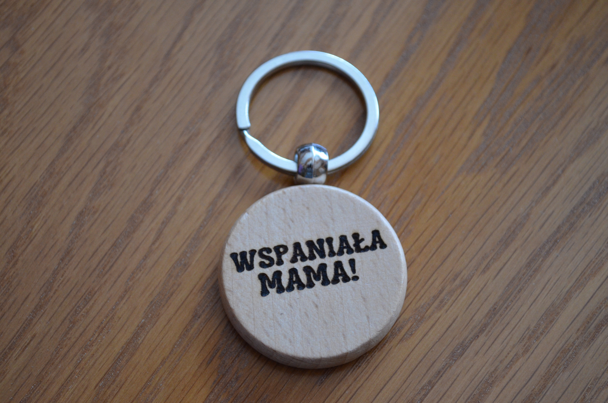 Drewniany breloczek z napisem 'Wspaniała Mama!' wykonany metodą CNC, leżący na drewnianym blacie.