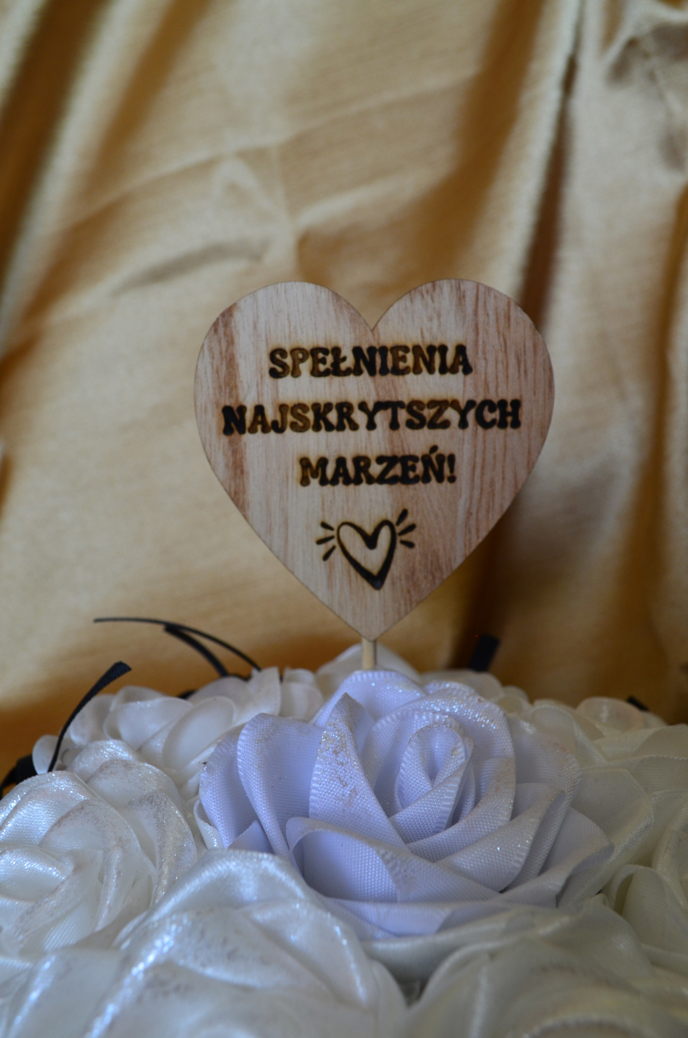 Drewniane serce z wypalonym laserowo napisem 'Spełnienia najskrytszych marzeń!' i symbolem serca, umieszczone w bukiecie białych, błyszczących, materiałowych róż na tle złotej tkaniny.