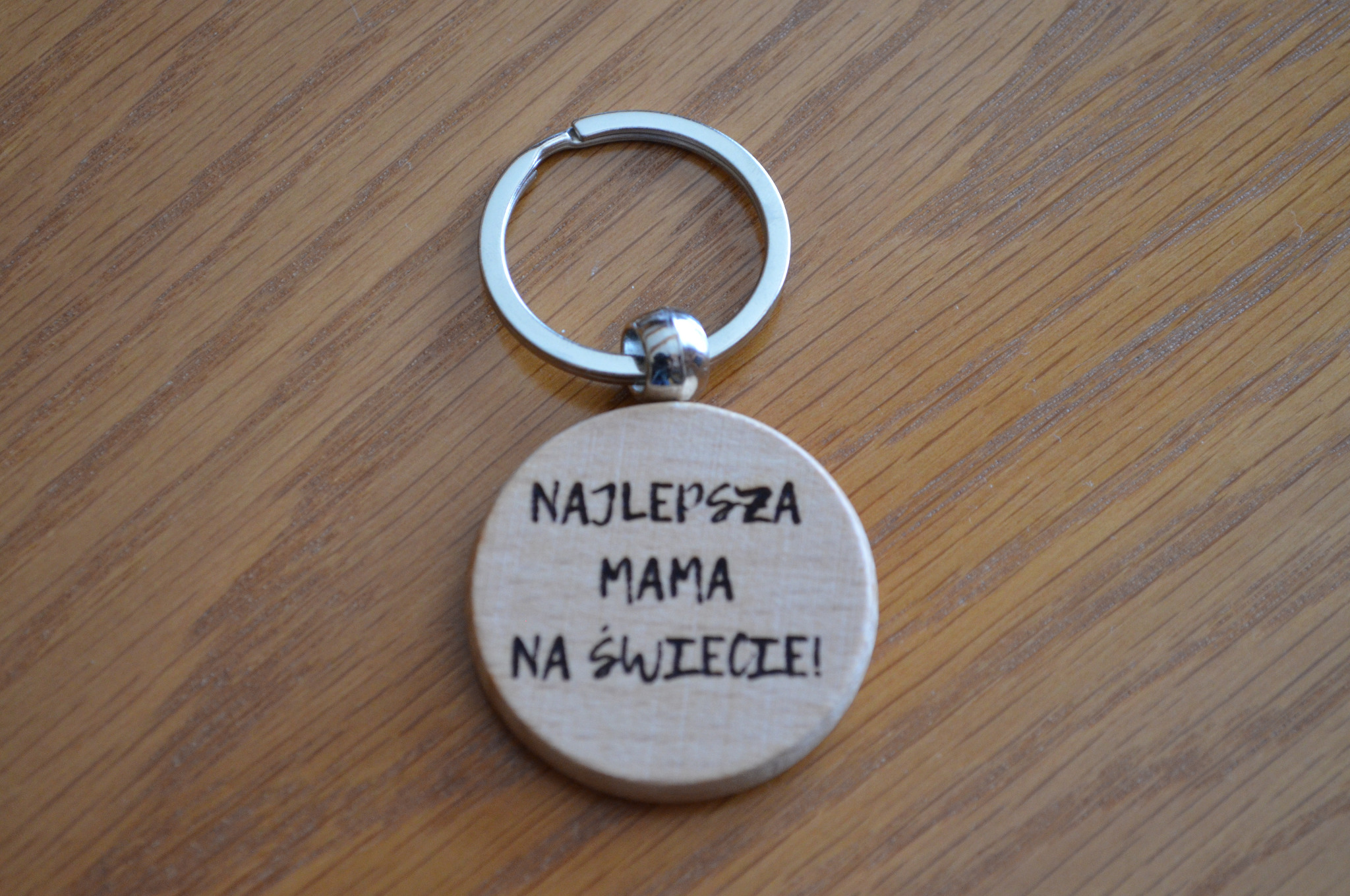 Drewniany breloczek z napisem 'Najlepsza Mama na świecie!' leżący na drewnianym blacie.