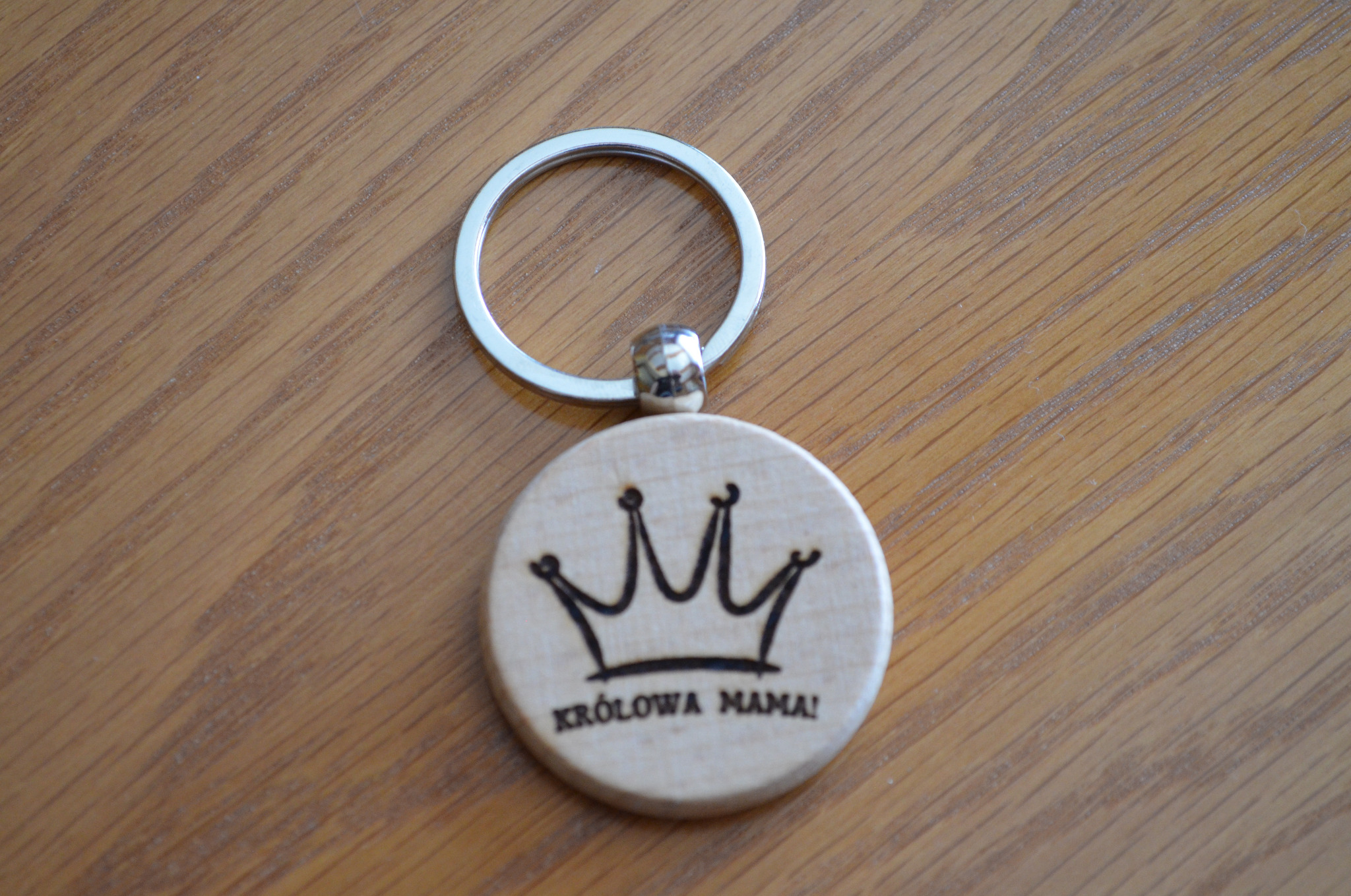 Drewniany breloczek z wypalonym napisem 'Królowa Mama!' i koroną, leżący na drewnianym blacie.