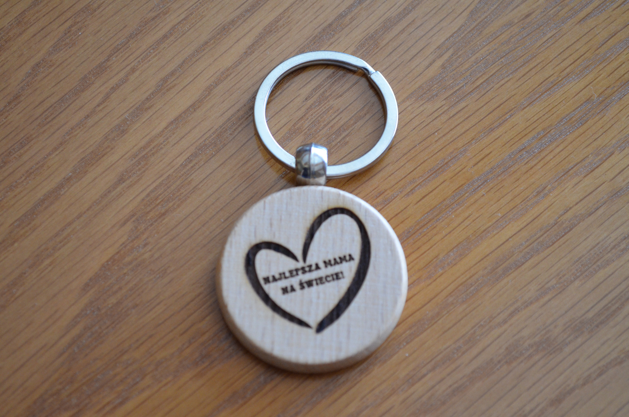 Drewniany brelok z wygrawerowanym laserowo napisem 'Najlepsza Mama na świecie' w kształcie serca, leżący na drewnianym blacie.
