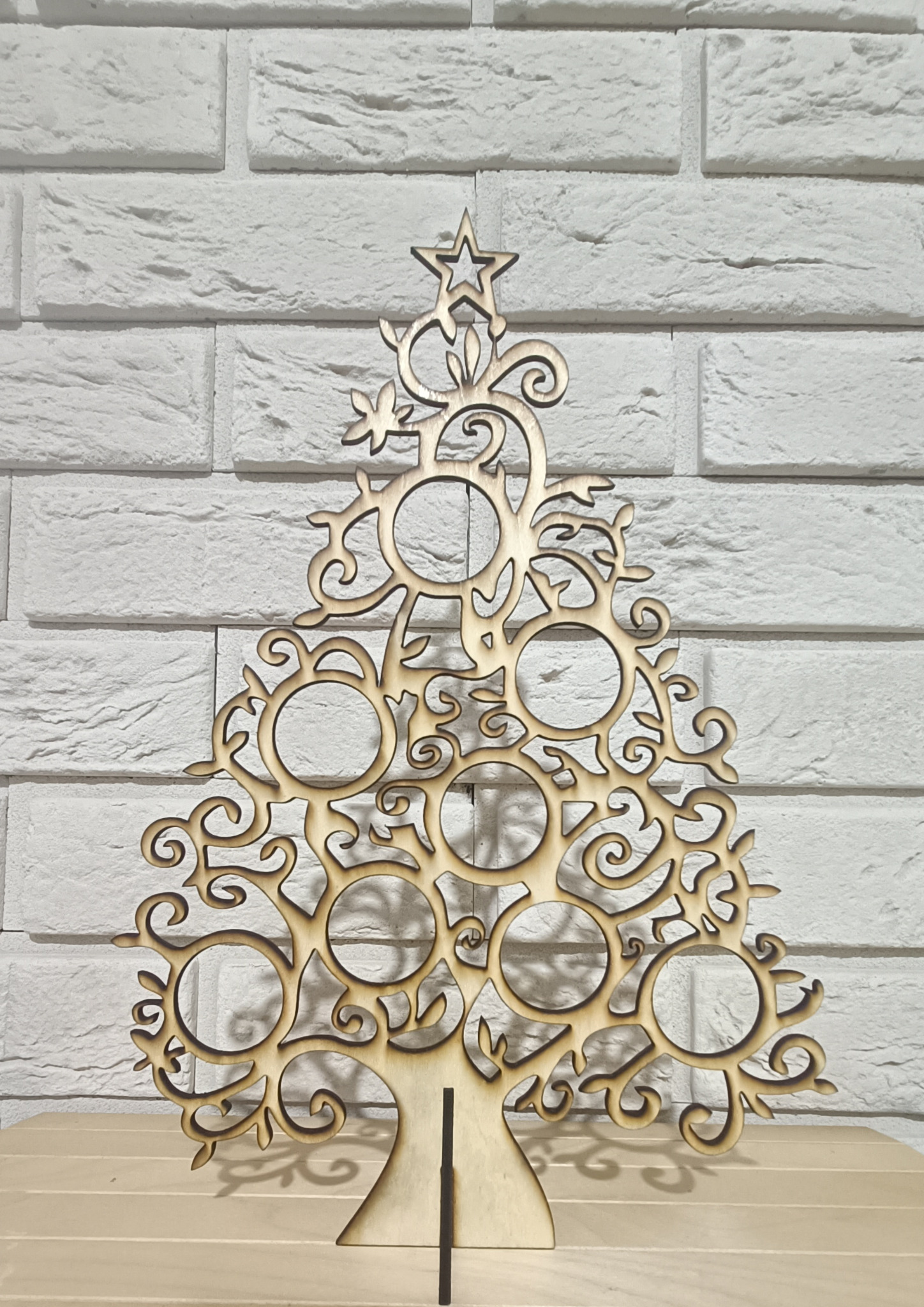 Dekoracyjna, ażurowa choinka z jasnego drewna, wycięta laserowo, na tle białej ceglanej ściany i drewnianego blatu.