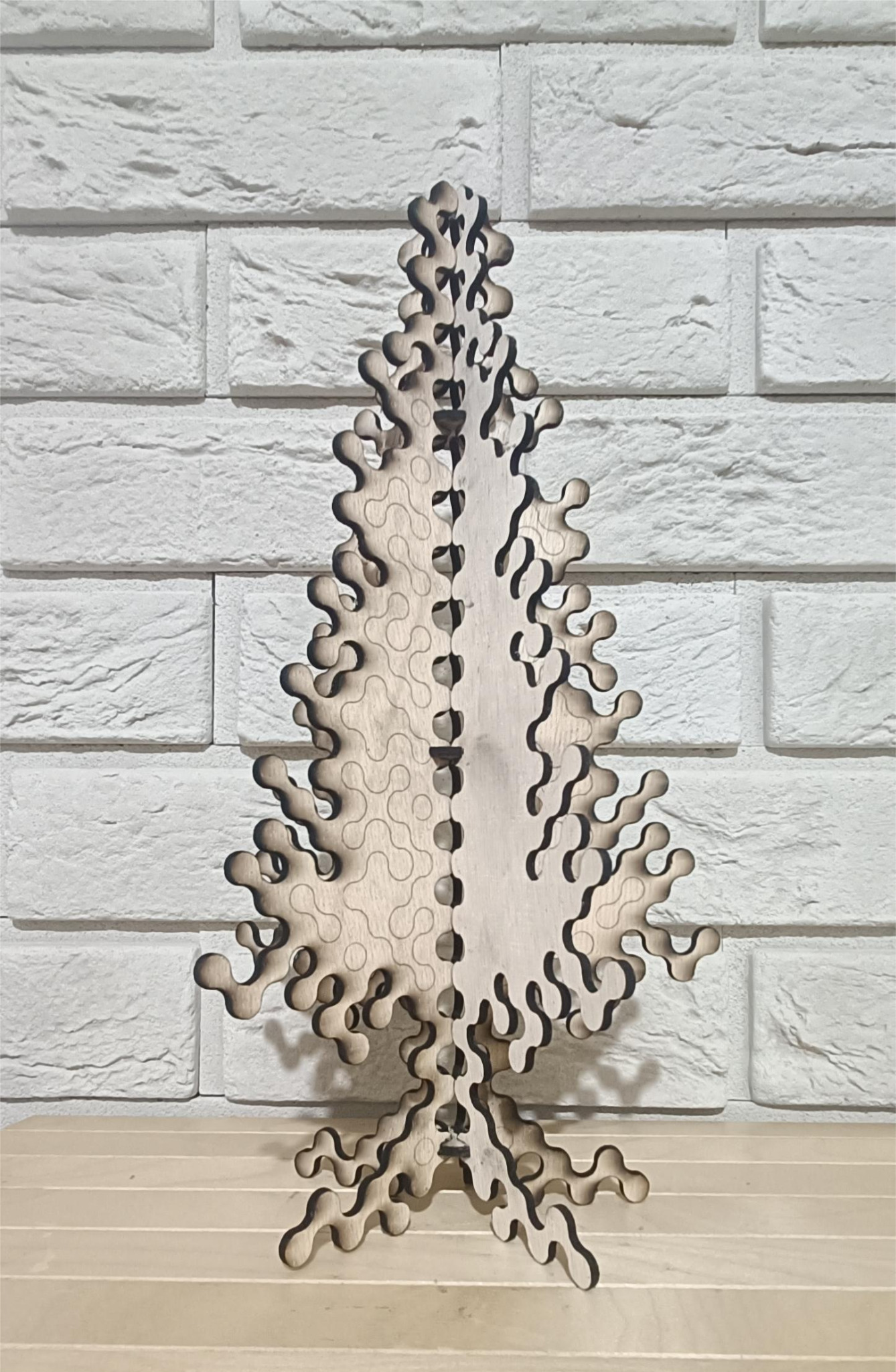 Drewniana, ażurowa choinka wycięta laserowo, stojąca na drewnianym blacie na tle białej ceglanej ściany.