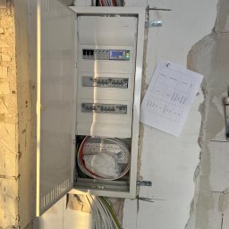 KP POWER - Otwarta skrzynka elektryczna z widocznymi bezpiecznikami, przewodami i schematem na ścianie w surowym stanie. Widoczne kable wchodzące do skrzynki.