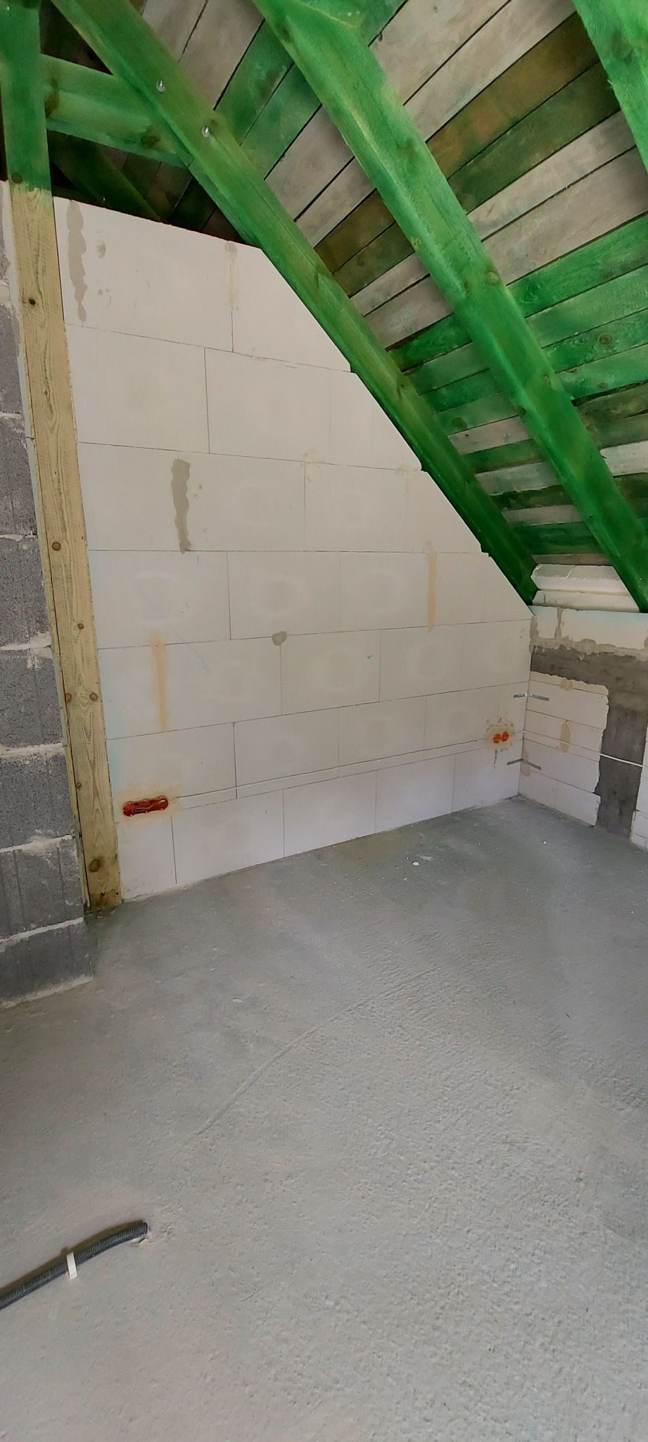 Wnętrze poddasza w trakcie prac wykończeniowych: ściana z białych bloczków, drewniana konstrukcja dachu pomalowana na zielono, widoczne punkty instalacji elektrycznej w ścianie.