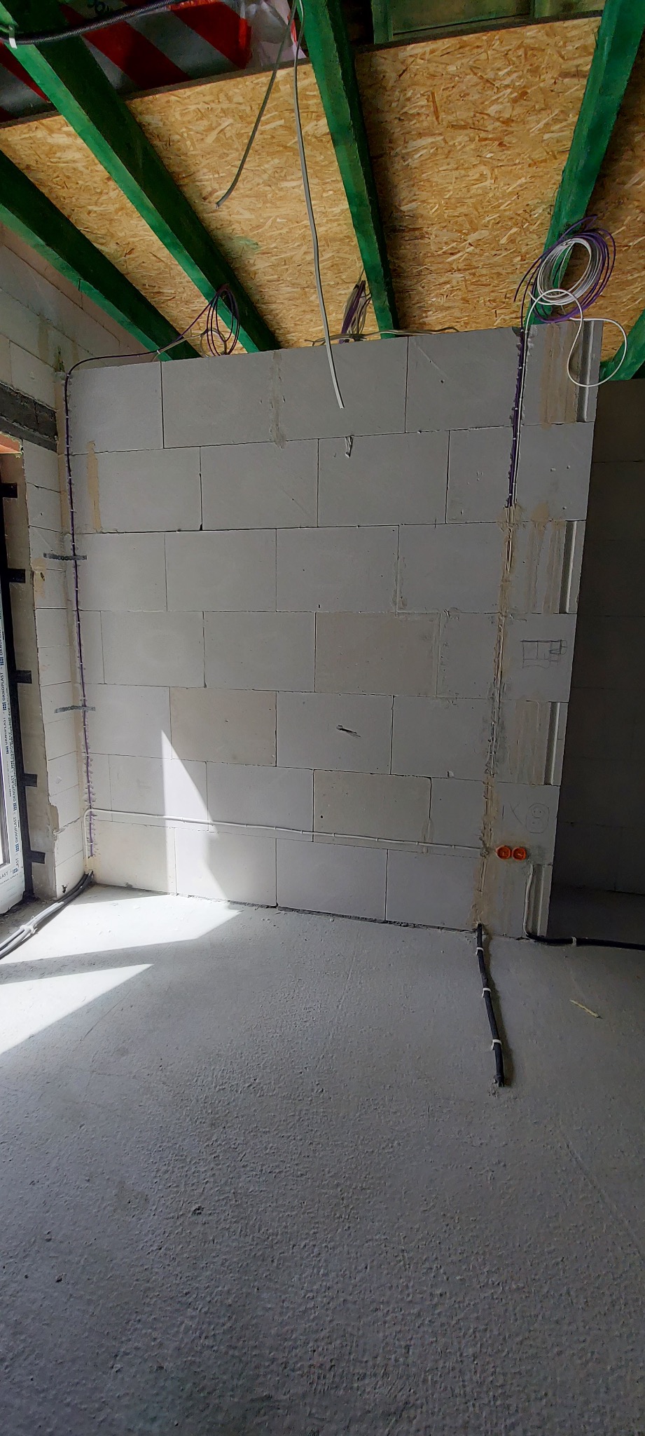 Surowy stan wnętrza z widoczną instalacją elektryczną: przewody prowadzone po ścianie z bloczków, puszki elektryczne, kable pod stropem z drewnianych belek.