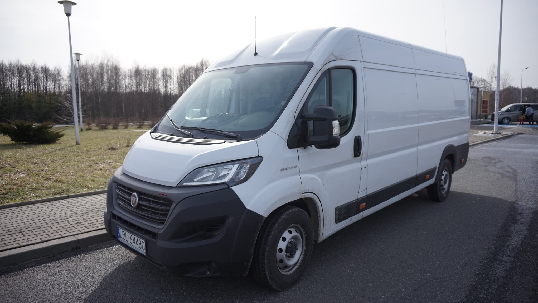 Biały samochód dostawczy Fiat Ducato zaparkowany na parkingu, widoczny z przodu pod kątem, z numerem rejestracyjnym LBL 64480, w tle drzewa bez liści i fragment budynku.