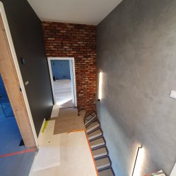 1. MARCUS BUDOWNICTWO I REMONTY WOJCIECH ZWARA - Klatka schodowa w trakcie remontu: ściana pokryta szarą gładzią, fragment ściany z cegły, schody zabezpieczone taśmą malarską i płytami, widoczny fragment pomalowanej na czarno ściany i wejście...