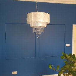 1. MARCUS BUDOWNICTWO I REMONTY WOJCIECH ZWARA - Ściana pomalowana na intensywny niebieski kolor z dekoracyjnymi ramkami, zwisająca biała lampa z kryształkami i fragment zielonej rośliny w rogu.