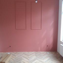 1. MARCUS BUDOWNICTWO I REMONTY WOJCIECH ZWARA - Ściana pomalowana na różowo z dwoma ozdobnymi prostokątnymi ramkami, podłoga wyłożona panelami w jodełkę, widoczne puszki elektryczne i kartonowe opakowanie na podłodze, surowa żarówka zwisająca...