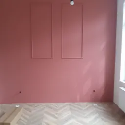 Ściana pomalowana na różowo z dwoma ozdobnymi prostokątnymi ramkami, podłoga wyłożona panelami w jodełkę, widoczne puszki elektryczne i kartonowe opakowanie na podłodze, surowa żarówka zwisająca...