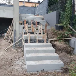 Betonowe schody zewnętrzne w trakcie budowy, z widocznym szalunkiem drewnianym, prowadzące w górę zbocza, z elementami krajobrazu w tle.