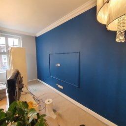 1. MARCUS BUDOWNICTWO I REMONTY WOJCIECH ZWARA - Ściana w kolorze głębokiego granatu z dekoracyjną ramą i ukrytymi gniazdkami elektrycznymi, biała listwa przypodłogowa i sufitowa, widoczne przygotowania do montażu telewizora, zakurzona podłoga...