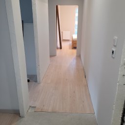 1. MARCUS BUDOWNICTWO I REMONTY WOJCIECH ZWARA - Korytarz w trakcie wykańczania: położone panele podłogowe w odcieniach beżu, świeżo pomalowane ściany na biało, widoczne niedokończone futryny drzwi.