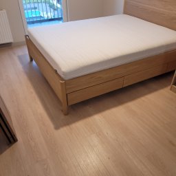 1. MARCUS BUDOWNICTWO I REMONTY WOJCIECH ZWARA - Jasna sypialnia z drewnianą podłogą i łóżkiem z materacem, balkon z widokiem na zieleń w tle. Minimalistyczny design.