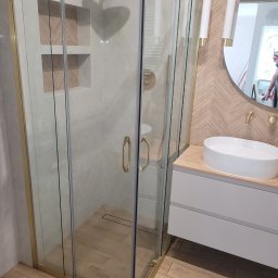 1. MARCUS BUDOWNICTWO I REMONTY WOJCIECH ZWARA - Nowoczesna łazienka: kabina prysznicowa ze złotymi profilami, umywalka nablatowa i lustro w drewnianej ramie. Widoczne oświetlenie punktowe i płytki imitujące drewno.