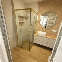 1. MARCUS BUDOWNICTWO I REMONTY WOJCIECH ZWARA - Nowoczesna łazienka z kabiną prysznicową w złotej ramie, okrągłym lustrem i umywalką nablatową na szafce z drewnianym blatem, wykończona płytkami imitującymi drewno.