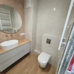 Jasna łazienka po remoncie: drewnopodobne płytki na podłodze i ścianie za umywalką, owalne lustro, podwieszana toaleta. Nowoczesny design.