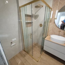 1. MARCUS BUDOWNICTWO I REMONTY WOJCIECH ZWARA - Elegancka łazienka z kabiną prysznicową w złotej ramie, drewnopodobne płytki na podłodze i ścianie, umywalka nablatowa i okrągłe lustro z oświetleniem.