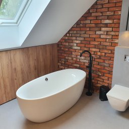 1. MARCUS BUDOWNICTWO I REMONTY WOJCIECH ZWARA - Nowoczesna łazienka na poddaszu z wolnostojącą wanną, czarną armaturą i ścianą z cegły, oświetlona światłem z okna dachowego. Drewniane panele i szara podłoga.