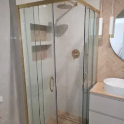 Nowoczesna kabina prysznicowa z złotymi detalami, wnęka z drewnianą mozaiką, umywalka nablatowa i lampy wiszące w wykończonej łazience. Jasne płytki, minimalistyczny design.