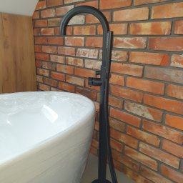 1. MARCUS BUDOWNICTWO I REMONTY WOJCIECH ZWARA - Nowoczesna, czarna bateria wannowa wolnostojąca na tle ściany z cegły. Wanna owinięta folią ochronną. Element wykończenia łazienki w stylu industrialnym.