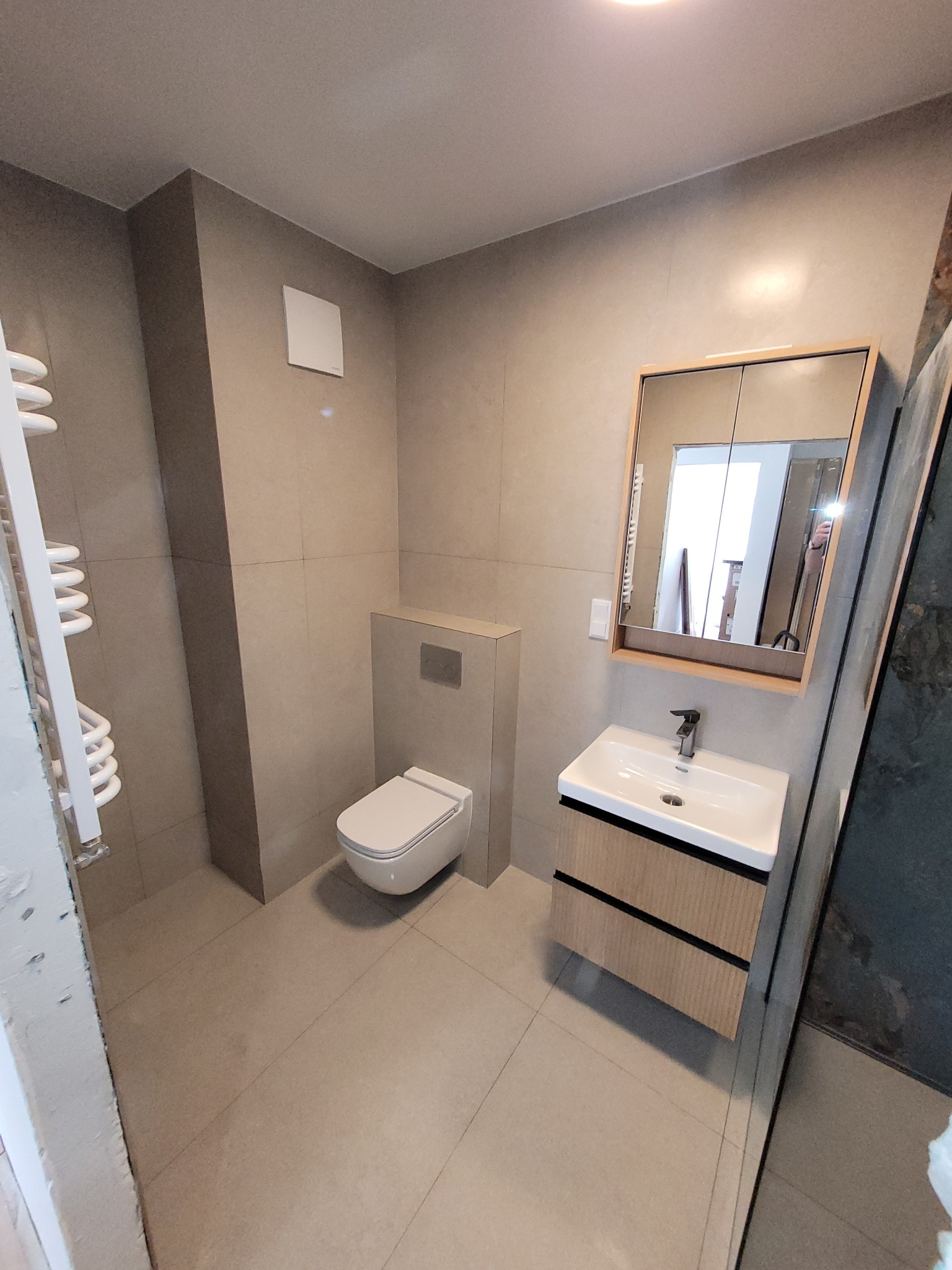 Wykończona łazienka w beżowych tonacjach z podwieszaną toaletą, umywalką z szafką i grzejnikiem drabinkowym. Minimalistyczny design, płytki w odcieniach beżu.