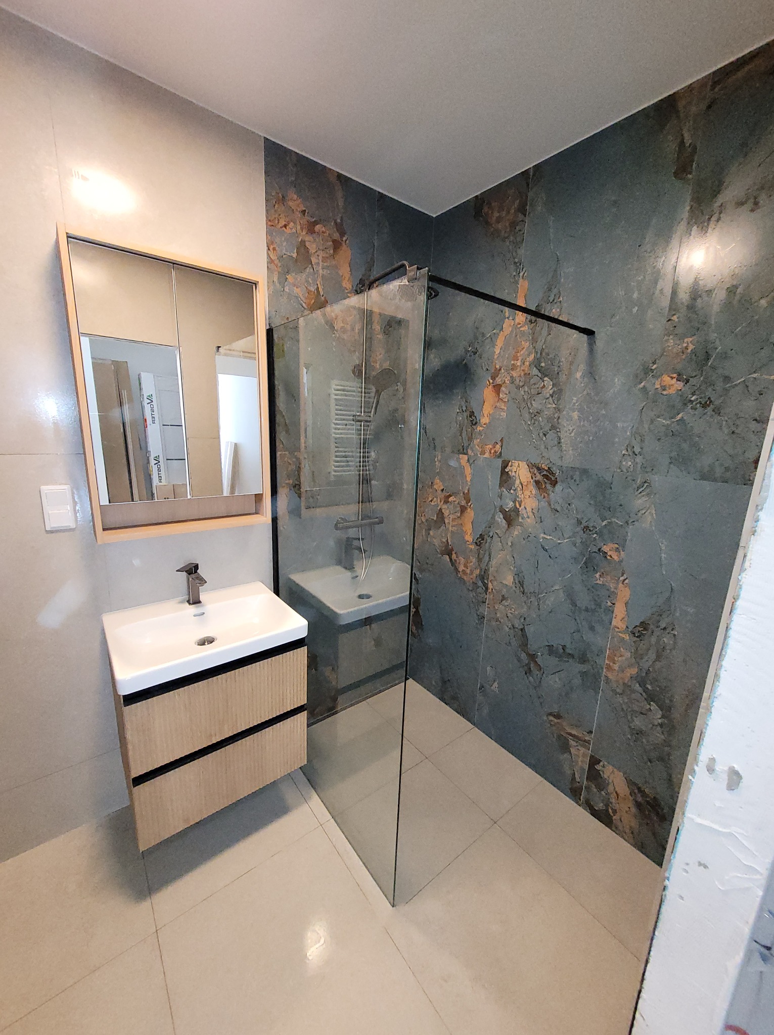 Nowoczesna łazienka po remoncie: szklana kabina prysznicowa, umywalka z szafką z drewnopodobnymi frontami, lustro z oświetleniem i płytki imitujące marmur.