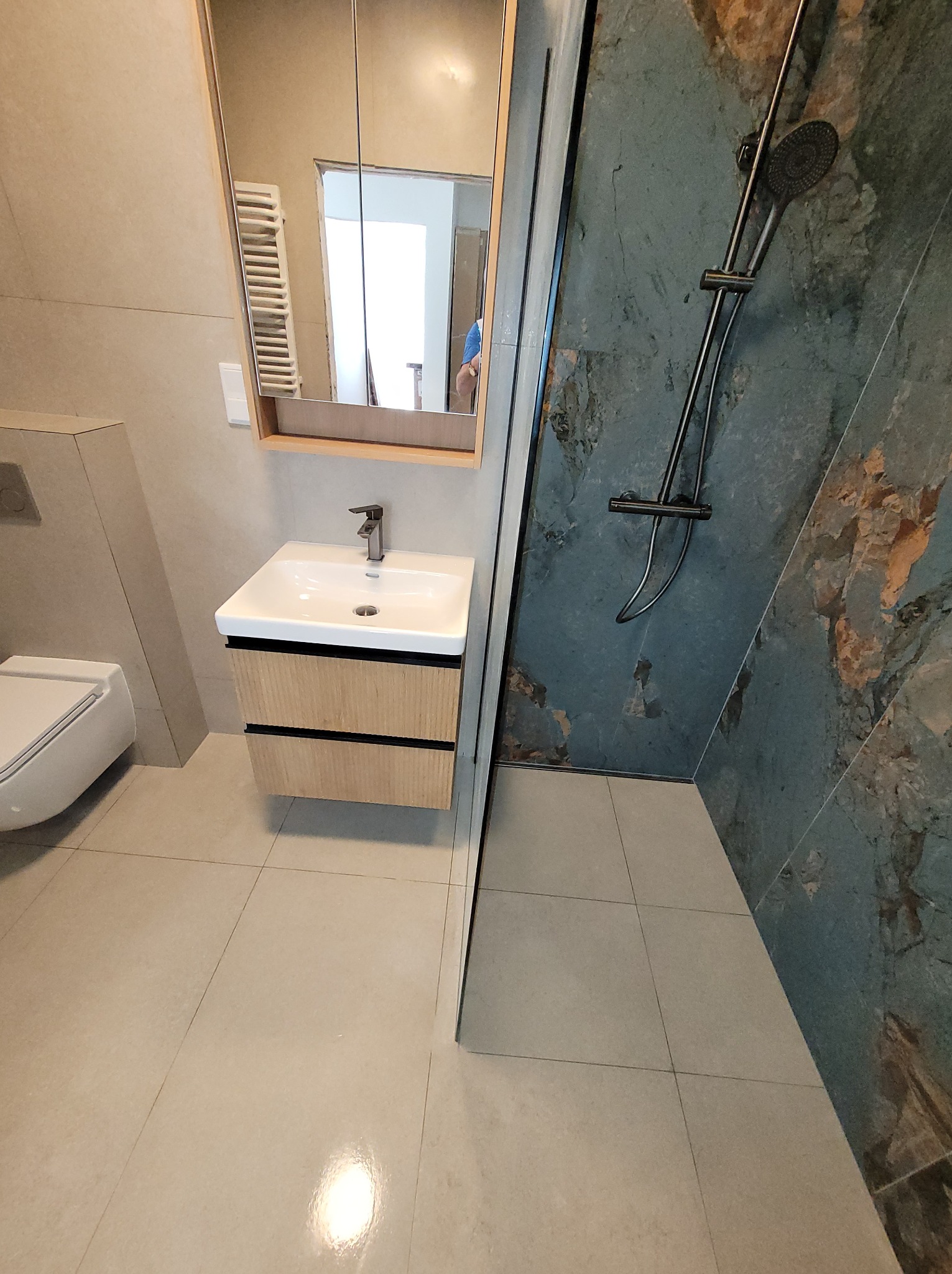 Nowoczesna łazienka z kabiną prysznicową, umywalką z szafką z jasnego drewna i lustrem, w którym odbija się grzejnik. Płytki w odcieniach szarości i beżu.