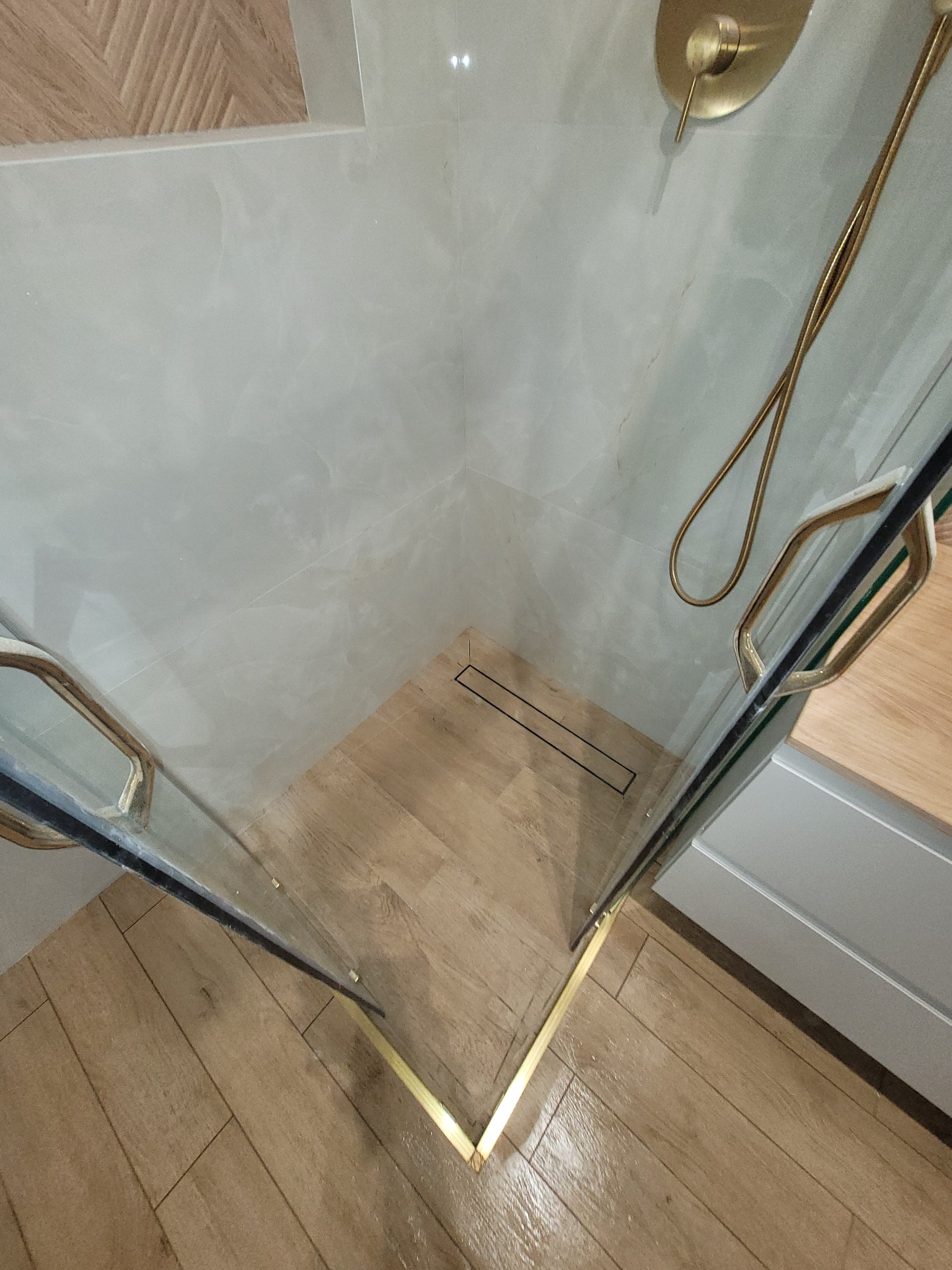 Nowoczesna kabina prysznicowa walk-in z złotymi detalami: armatura, listwy wykończeniowe. Płytki imitujące drewno na podłodze, jasne płytki na ścianach.